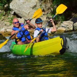 canoe jaune et vert avec un homme et un jeune adolescent avec des gilets bleus et des pagaies jaunes, l'un est chauve, l'autre à une casquette noire et bleue, ils passent un rapide sur la rivière Ardèche