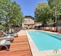 une piscine remplit devant une maison autour une terrasse en bois avec des chaises longues avec des serviettes roulées dessus, logo gîte de france , des murets en pierre